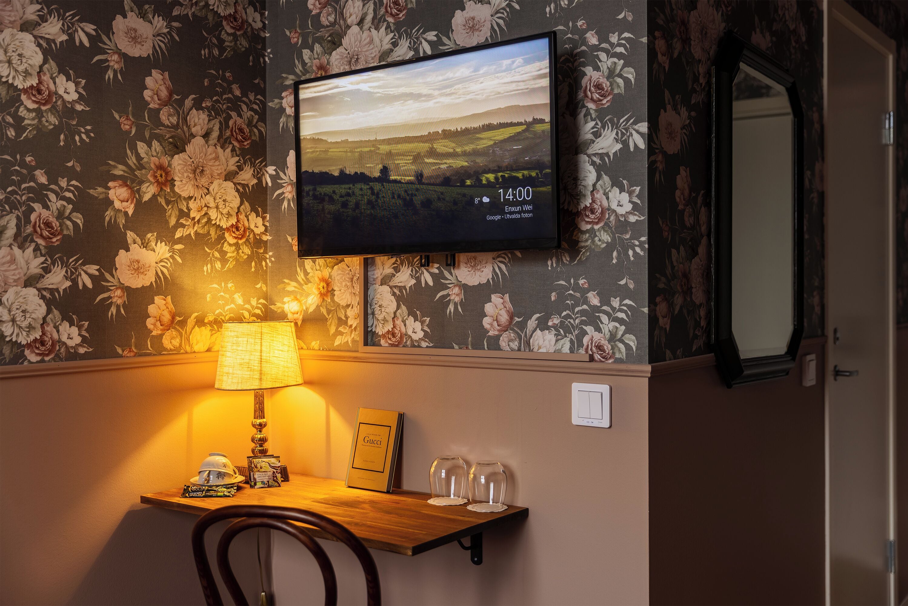 Una habitación con una televisión mostrando un paisaje, una mesa con una lámpara, una silla y un cuadro enmarcado.