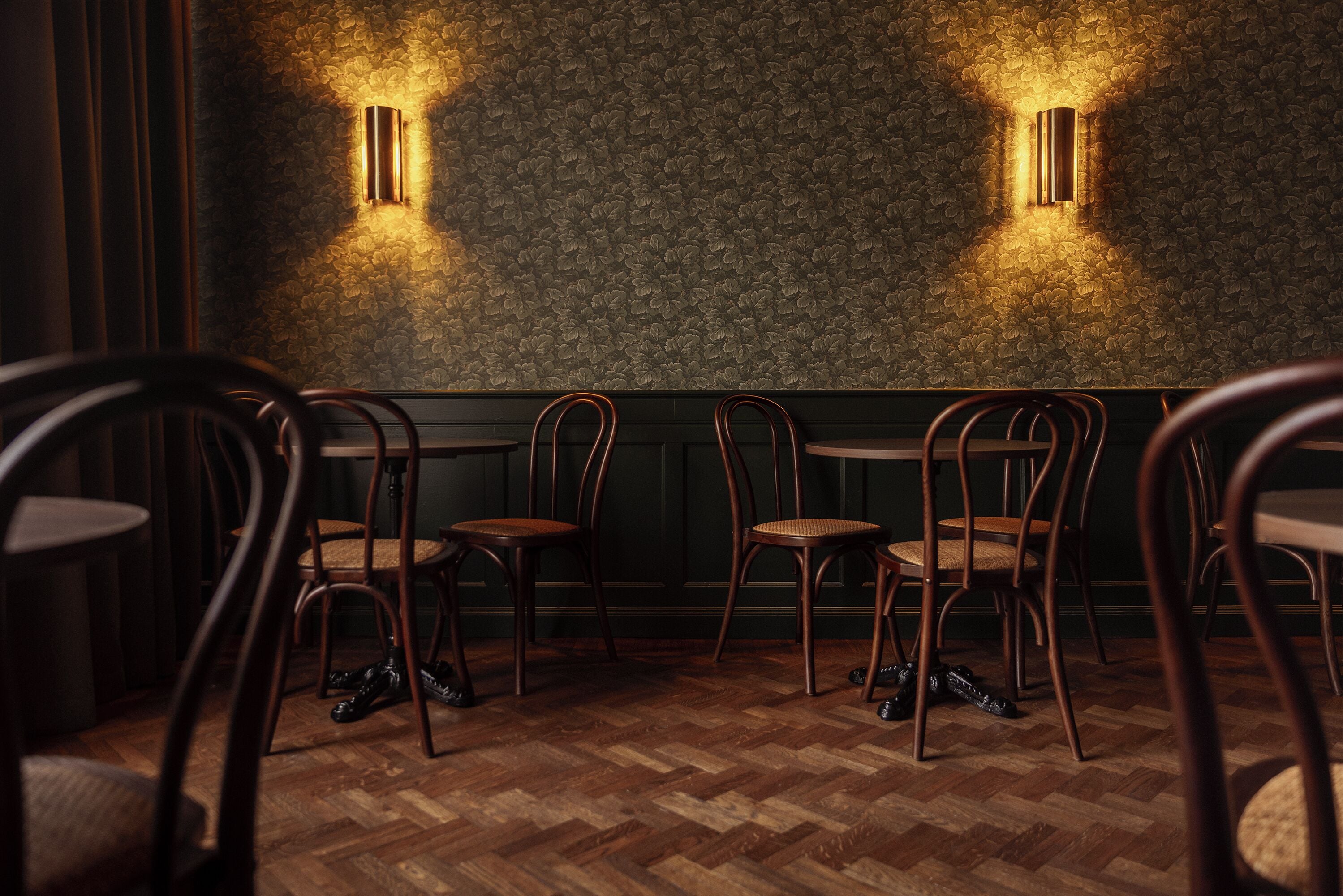 Una habitación tenuemente iluminada con sillas y mesas de madera, un papel tapiz estampado e iluminación en la pared.
