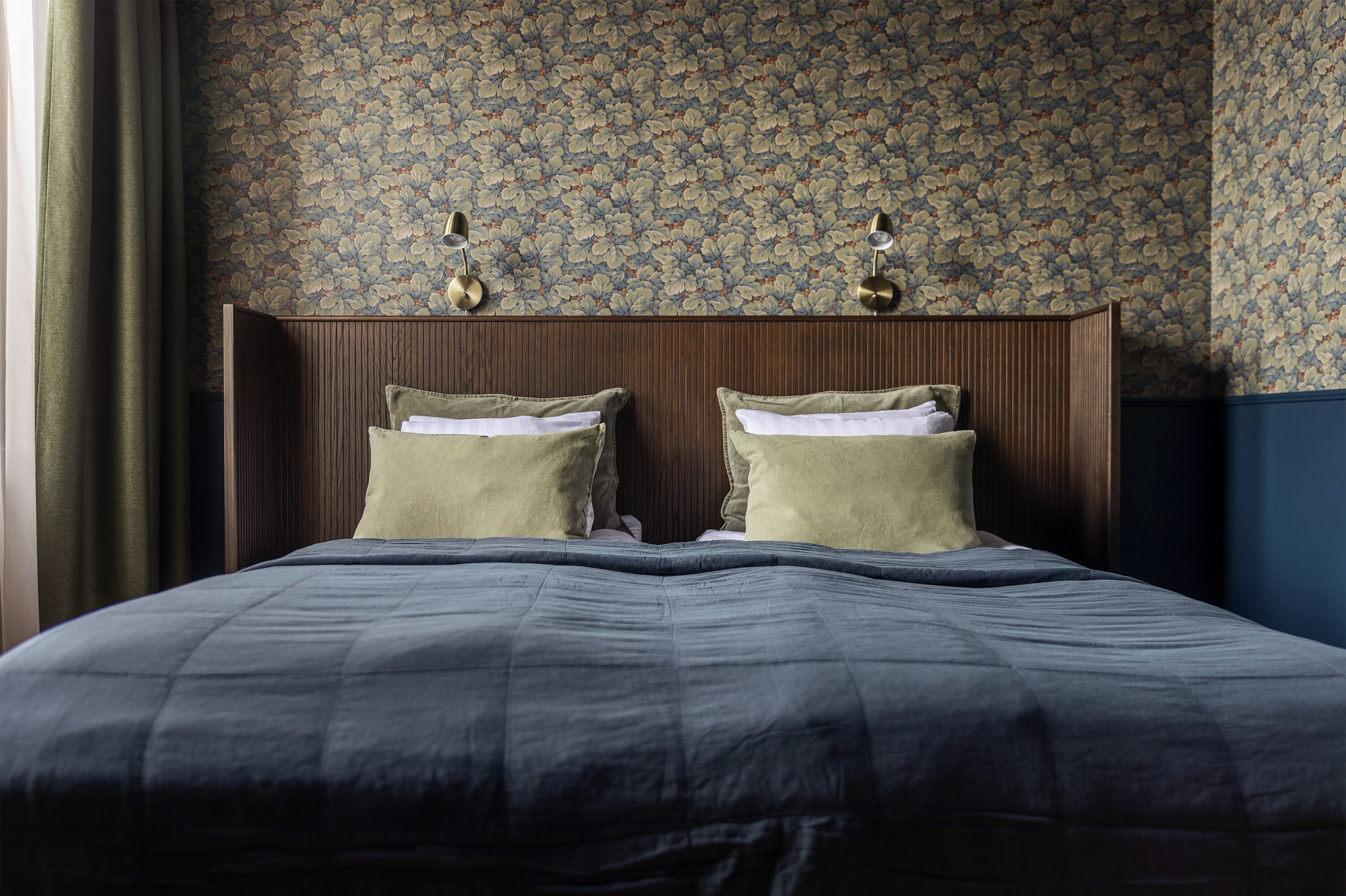 A neatly made bed with a dark blue comforter and two pillows, a wooden headboard, and floral wallpaper.
