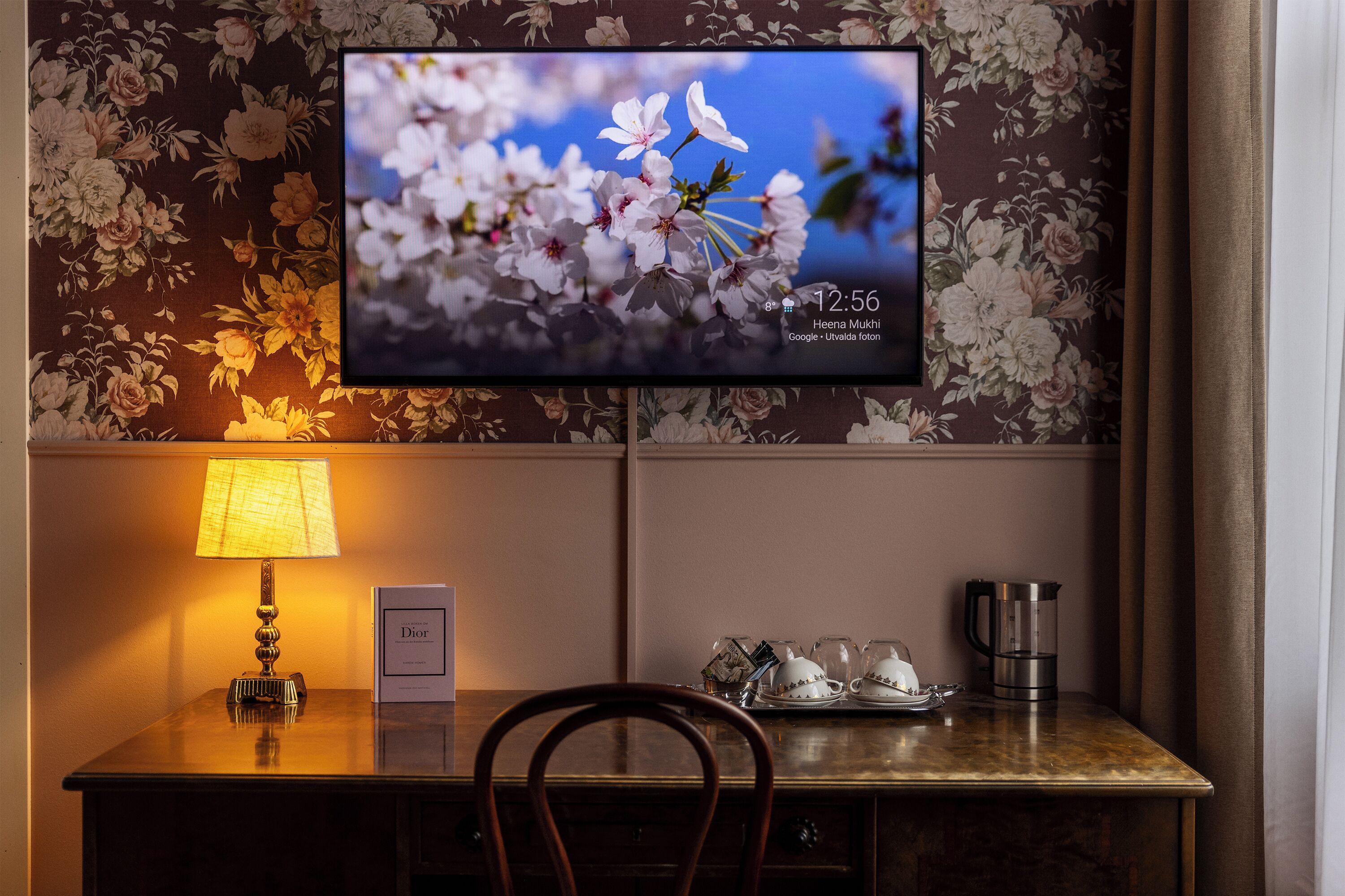 A hotel room with a floral wallpaper, a television displaying a floral image, a bedside table with a lamp, a sign, and a coffee maker.