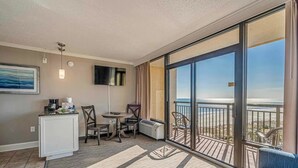 Quarto de hotel com varanda, área de refeições com cadeiras, TV fixada na parede e vista para a praia.