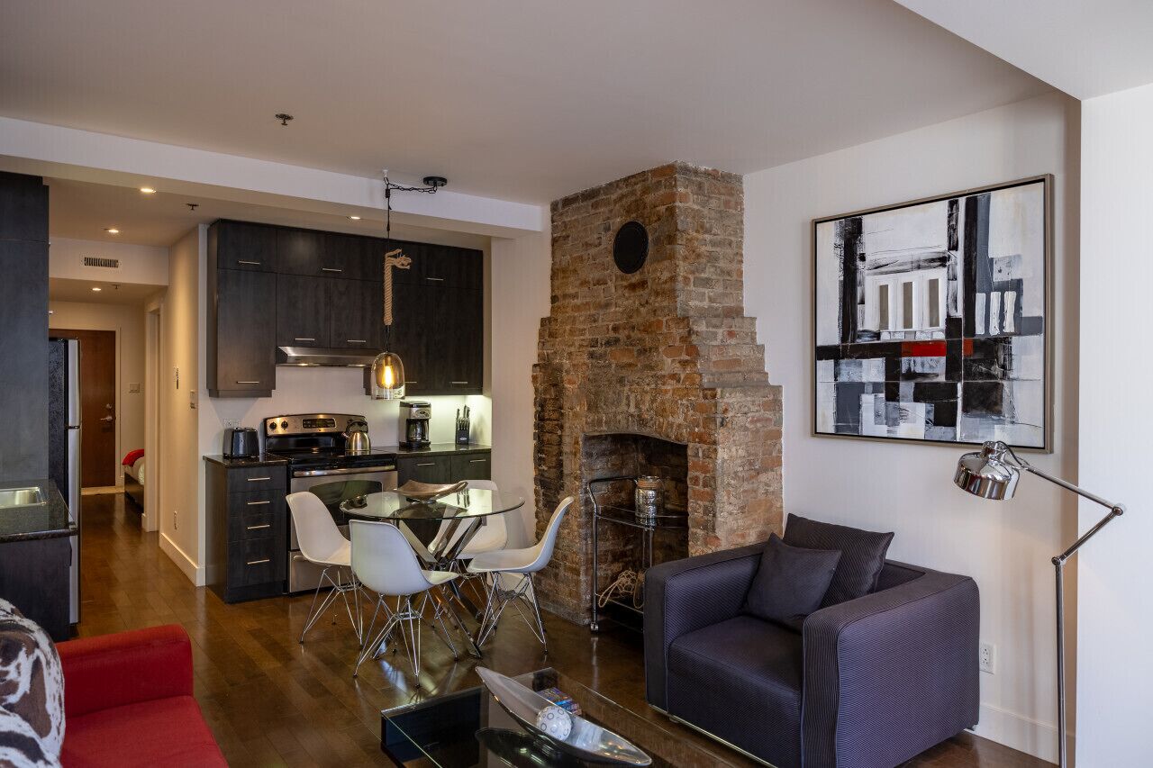 Uma sala de estar moderna com lareira, uma área de jantar com mesa de vidro e uma cozinha com armários escuros e eletrodomésticos de aço inoxidável.
