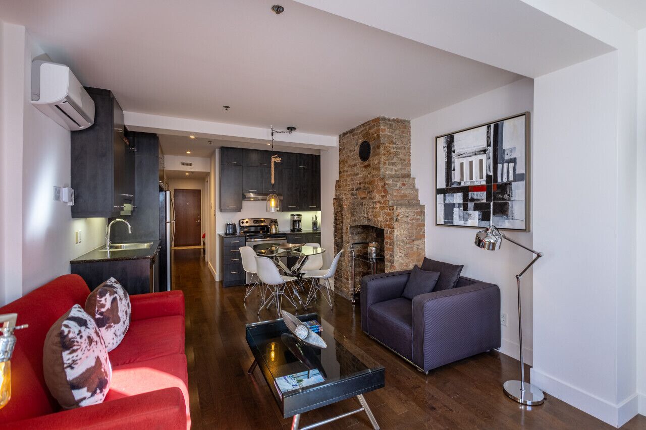 Una sala moderna con un sofá rojo, un sofá gris oscuro, una mesa de centro y una chimenea. Se ve una cocina al fondo con armarios oscuros y una pared de ladrillo.