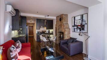 Una sala moderna con un sofá rojo, un sofá gris oscuro, una mesa de centro y una chimenea. Se ve una cocina al fondo con armarios oscuros y una pared de ladrillo.