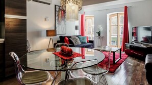 A modern living room with a glass dining table, clear chairs, a sofa with red and grey cushions, a large chandelier, and a television. - Les Immeubles Charlevoix - Le Quai St-André 1166 (Québec City)