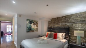 A bedroom with a stone wall, a bed with white bedding, red and grey pillows, a nightstand with a lamp, and a painting on the wall. - Les Immeubles Charlevoix - Le Quai St-André 1166 (Québec City)