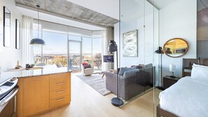 Ein modernes Hotelzimmer mit Küchenzeile, einer Sitzecke, einem Bett und einem großen Fenster mit Blick auf die Stadt.
