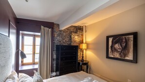 A hotel room with a bed, a dresser, a lamp, a painting, and a stone wall. - 1002 · Les Immeubles Charlevoix - 1002 (Québec)