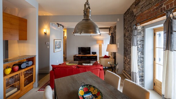 A modern living room with a red sofa, a dining table with chairs, a TV, and a stone wall. - 1002 · Les Immeubles Charlevoix - 1002 (Québec)