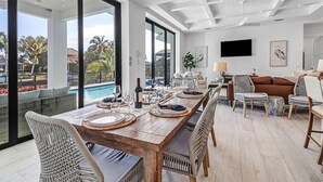 A spacious dining area with a view of a pool and palm trees, a comfortable seating area with wicker furniture, and a flat-screen TV mounted on the wall.