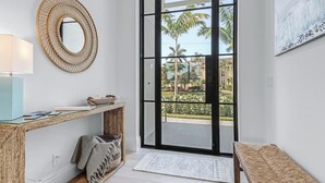 A modern entryway with a wicker console table, a mirror, a bench, and a view of palm trees through a glass door.