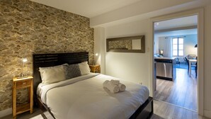 A bedroom with a stone wall, a bed with white linens, a wooden nightstand, and a modern bathroom visible through an open door.