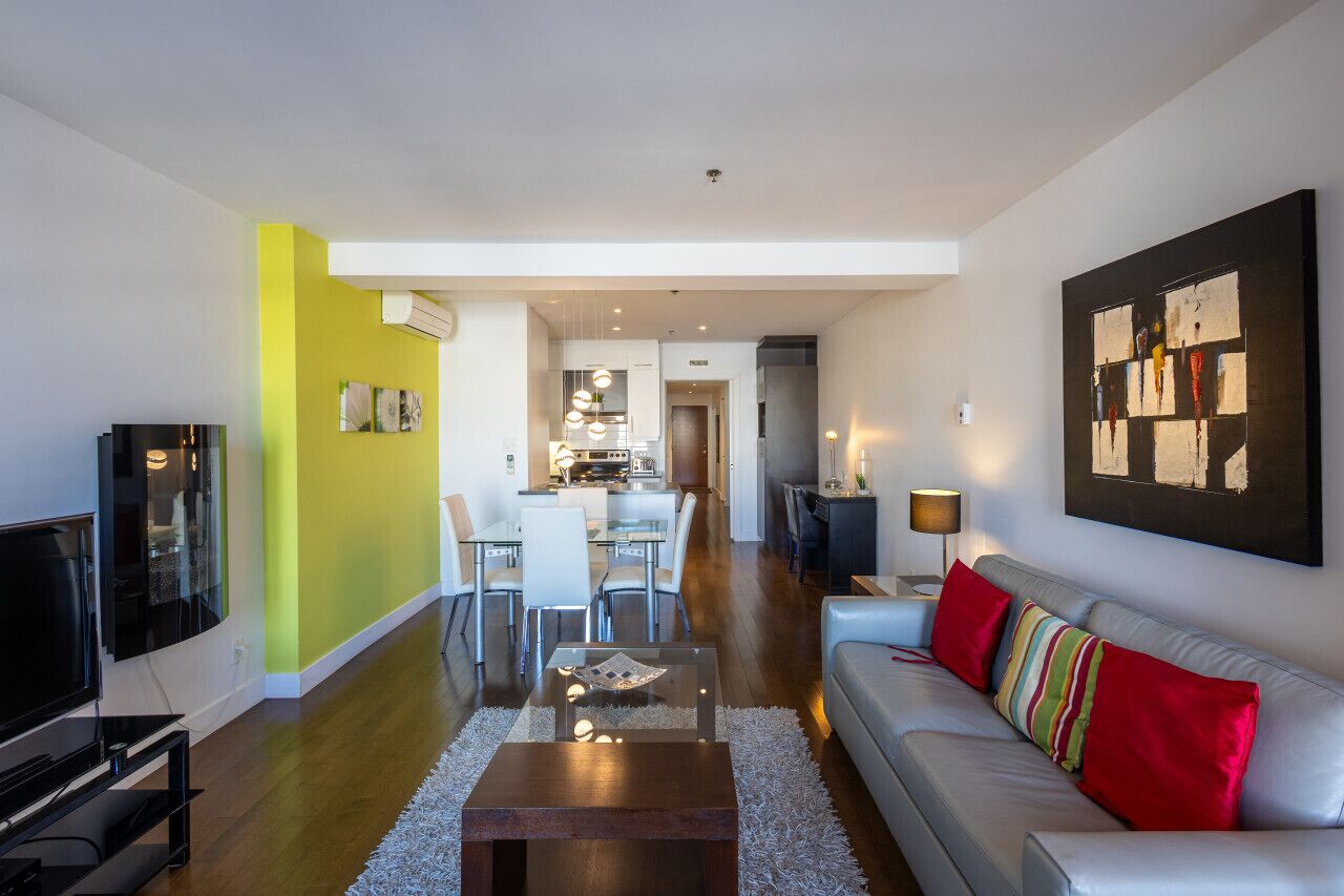 Ein modernes Wohnzimmer mit Sofa, Couchtisch und Fernseher. Es gibt einen Essbereich mit Stühlen und einem Tisch. Der Raum hat eine leuchtend gelbe Akzentwand und ein großes abstraktes Gemälde an der Wand.