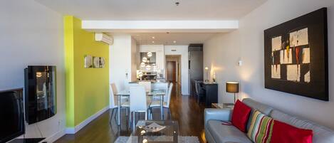 Ein modernes Wohnzimmer mit Sofa, Couchtisch und Fernseher. Es gibt einen Essbereich mit Stühlen und einem Tisch. Der Raum hat eine leuchtend gelbe Akzentwand und ein großes abstraktes Gemälde an der Wand.