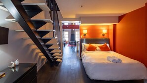Habitación de hotel con cama, mesita de noche, escritorio, silla, escalera y vista al comedor.