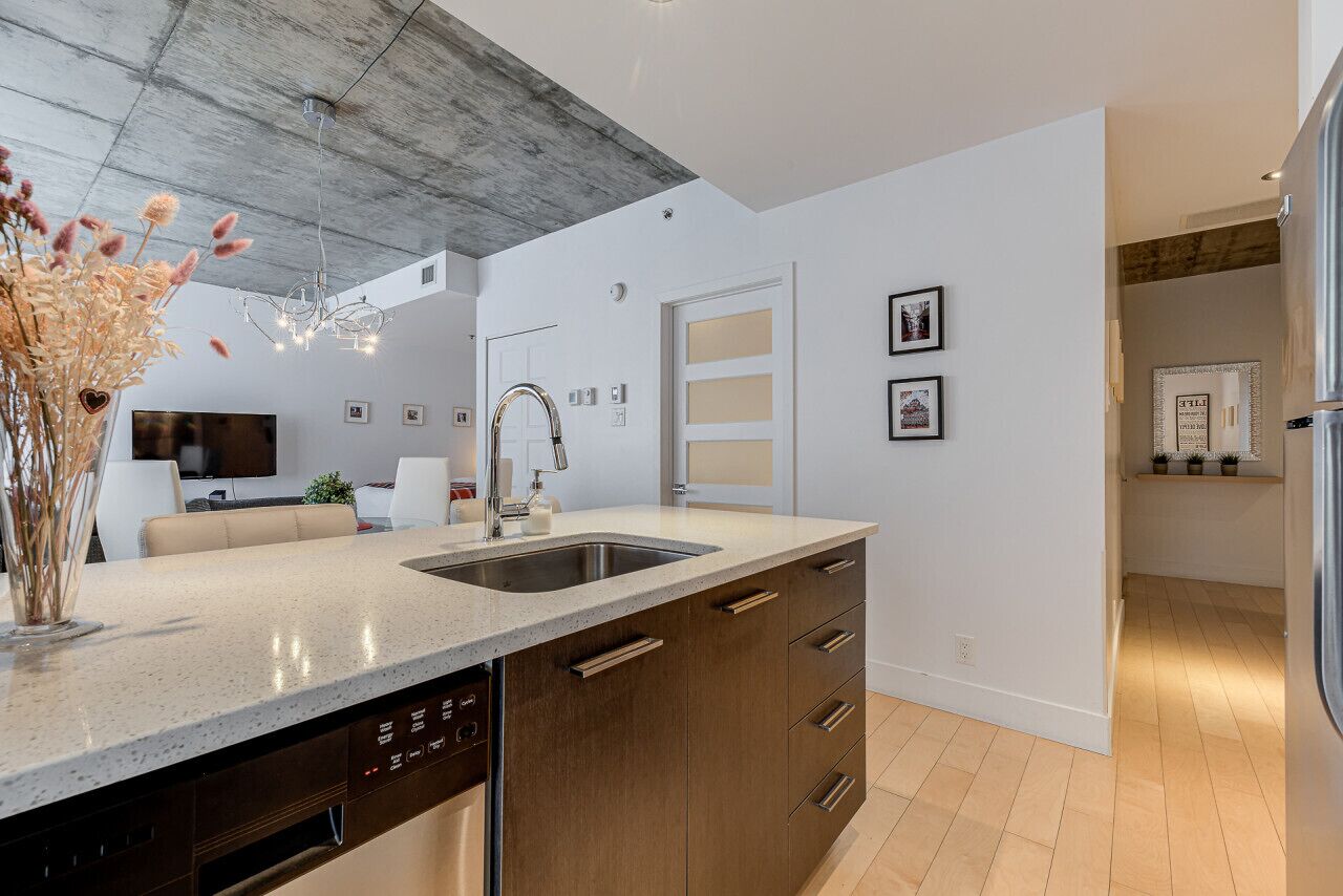 A modern kitchen with a dishwasher, stainless steel sink, and wooden cabinets. There is a dining area with a sofa and a TV.