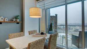 A modern dining area with a wooden table and woven chairs, large windows offering a city view, and a hanging light fixture. - Les Immeubles Charlevoix - 760214 (Ville de Québec)