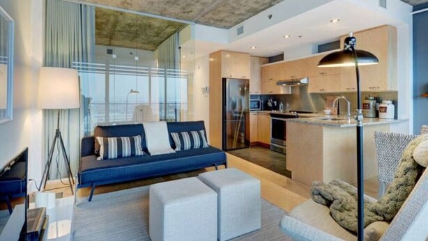 A modern living room with a sofa, ottoman, and a lamp. There is a kitchen area with a refrigerator, oven, and sink. - Les Immeubles Charlevoix - 760214 (Ville de Québec)