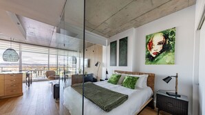 A modern bedroom with a large bed, a glass partition, a city view, and a painting on the wall.