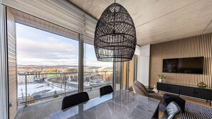 A modern living room with a large dining table, a black woven pendant light, a flat-screen TV mounted on the wall, and a view of a cityscape through large windows.