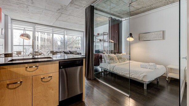 Une cuisine moderne avec des meubles en bois et un lave-vaisselle, ainsi qu’une cloison en verre qui sépare la cuisine d’une chambre à coucher équipée d’un lit et d’une table de chevet.
