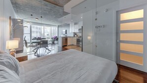 Une chambre moderne avec un grand lit, une porte en verre dotée de panneaux lumineux, et une vue sur un coin repas et une cuisine en arrière-plan.