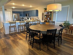 Un coin repas avec une table ronde en bois entourée de chaises, une cuisine en arrière-plan et une bibliothèque agrémentée de plantes.