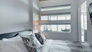 A modern hotel room with a large bed, a flat-screen TV, and multiple windows providing natural light. - Les Immeubles Charlevoix - 760431 (Ville de Québec)