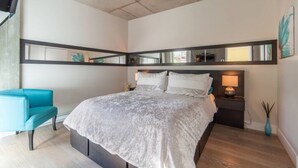 Un dormitorio moderno con una cama, una mesita de noche, un sillón azul y un espejo sobre la cama.