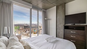 A hotel room with a large bed, a TV, and a view of a cityscape through large windows. - Les Immeubles Charlevoix - 760415 (Ville de Québec)