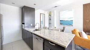 Una cocina moderna con encimera de granito, electrodomésticos de acero inoxidable y una zona de comedor con una silla amarilla y un cuadro del océano.
