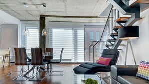 A modern living room with a black leather sofa, dining area with metal chairs, a staircase with a black railing, and a large window with blinds. - Les Immeubles Charlevoix - 760506 (Ville de Québec)