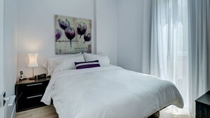 Un letto rifatto con lenzuola bianche, un cuscino viola e un quadro appeso al muro raffigurante fiori viola.