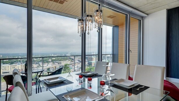 A modern dining area with a glass table, white chairs, and a city view. - Les Immeubles Charlevoix - 760514 (Ville de Québec)