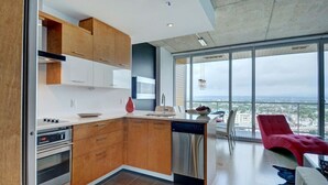 A modern kitchen with wooden cabinets, a dishwasher, and a large window overlooking a cityscape. - Les Immeubles Charlevoix - 760514 (Ville de Québec)