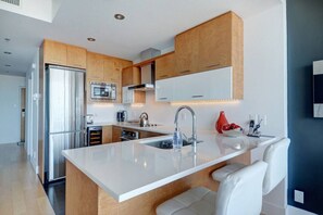 A modern kitchen with wooden cabinets, a stainless steel refrigerator, a microwave, and a large island with a sink. - Les Immeubles Charlevoix - 760514 (Ville de Québec)