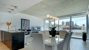 A modern kitchen with a central island, dining area with white chairs, and large windows offering a city view.