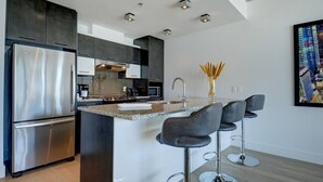 A modern kitchen with a stainless steel refrigerator, dark cabinets, a central island with bar stools, and recessed ceiling lighting.