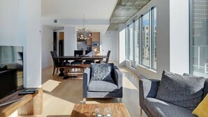 Een moderne woonkamer met een grote televisie, comfortabele zitplaatsen, een eethoek en grote ramen met uitzicht op de stad.