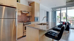 Eine moderne Küche mit einem Edelstahl-Kühlschrank, einem Herd und einer Mikrowelle. Eine große Kochinsel mit Spüle und einem schwarzen Barhocker. Die Küche verfügt über Holzschränke und eine geflieste Rückwand.