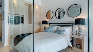 Une chambre moderne dotée d’un dressing fermé par des parois de verre, d’un lit avec une literie blanche, de deux lampes de chevet et de trois miroirs ronds accrochés au mur.