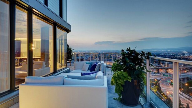 Un balcón moderno con un sofá blanco y cojines, una planta grande en maceta y vistas a la ciudad al atardecer.