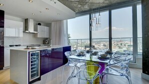 Una cocina moderna con una isla grande, una zona de comedor con sillas transparentes y vistas a la ciudad a través de amplios ventanales.