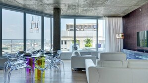 Una sala de estar moderna con una mesa de comedor de vidrio, sillas transparentes, un sofá blanco y un amplio ventanal con vistas a la ciudad.