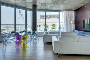 A modern living room with a glass dining table, clear chairs, a white sofa, and a large window offering a city view. - Les Immeubles Charlevoix - 760707 (Ville de Québec)