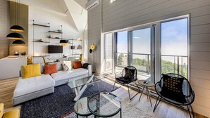 Una sala de estar moderna con sofá, mesa de centro y sillones, ventanas amplias con vistas, y televisor montado en la pared.