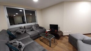 Un salon moderne avec un canapé d’angle gris, une petite table basse et un téléviseur posé sur un meuble.