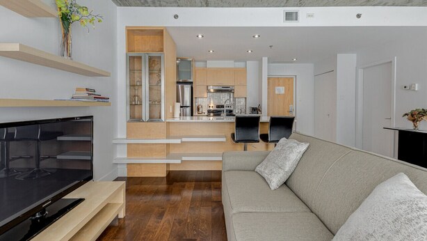 Una sala de estar moderna con una televisión de pantalla plana, un sofá gris, estanterías de madera y una cocina al fondo.