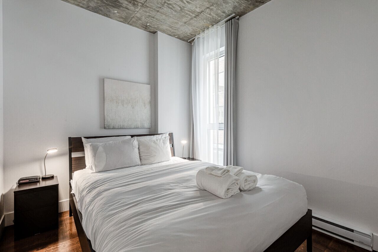 Une chambre d’hôtel moderne avec un grand lit, une table de chevet avec une lampe et une vue sur la ville à travers des rideaux transparents.