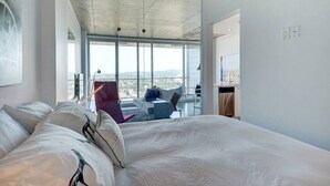 Une chambre d’hôtel moderne dotée d’un grand lit, offrant une vue sur la ville grâce à de larges fenêtres, et disposant d’un balcon avec une chaise et une table.